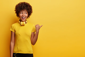 Wall Mural - Waist up shot of African American hipster girl being amused by listening music, has modern headphones on neck, points aside, shows way to electronic shop, has cheerful expression, isolated on yellow