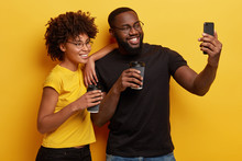 Lovely Dark Skinned African Couple Take Selfie With Modern Smart Phone, Drink Coffee To Go, Smile Broadly At Camera, Enjoy Spare Time, Stands Closely To Each Other Against Yellow Background.