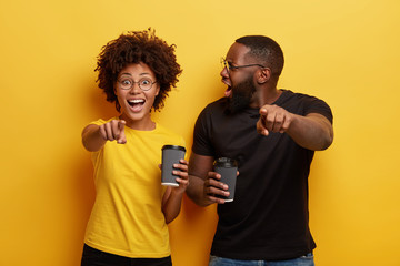 Wall Mural - Happy dark skinned woman and her boyfriend being emotional, surprised, point at camera, laugh at something funny, drink takeaway coffee, enjoy free time together, react on something astonishing