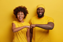 Studio Shot Of Overjoyed Satisfied Woman And Man With Dark Skin, Give Fist Bump, Agree To Work Together, Have Success In Project Work, Wear Yellow Attire, Pose In Studio. Cooperation Concept