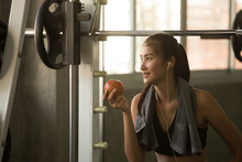 Asian Fitness Woman With Towel And Healthy In Gym Sitting On Weight Lifting And Resting After Workout Or Excercise And Looking Outside Listening Music And Holding Red Apple For Healthy And Smile