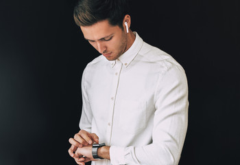 Studio portrait of smart young man wearing white shirt, looking at smartwatch posing over black background hurry up for meeting. People, business and technology concept