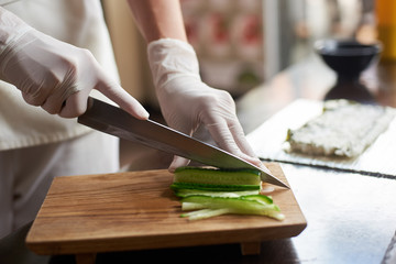 Close up view of process of preparing delicious rolling sushi in restaurant. Female hands in disposable gloves slicing cucumber on wooden board.