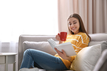 Canvas Print - Young woman with cup of coffee reading book on sofa at home