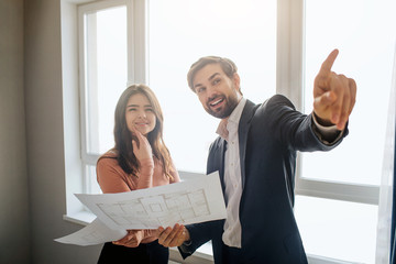 Couple buy or rent apartment together. Young man hold plan in hand and point up forward. Stand together at window. Smile and dream. Daylight.