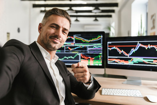 Photo of handsome man taking selfie while working in office on computer with graphics and charts at screen