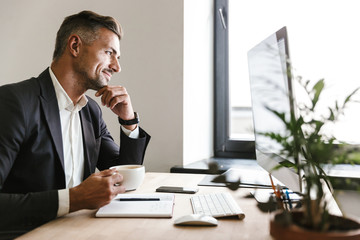 Fototapete - Image of unshaved businessman drinking coffee while working on computer in office