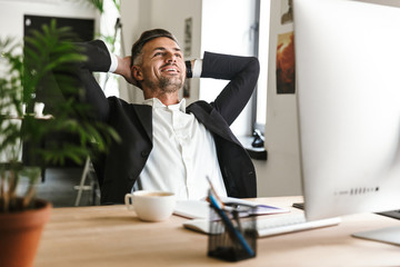 Fototapete - Photo of beautiful businessman smiling while sitting at desk and working on computer in office