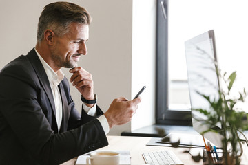 Fototapete - Portrait of attractive businessman using mobile phone while working on computer in office