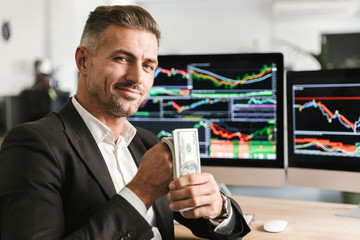 Fototapete - Image of rich businessman holding pack of money while working in office with graphics and charts on computer