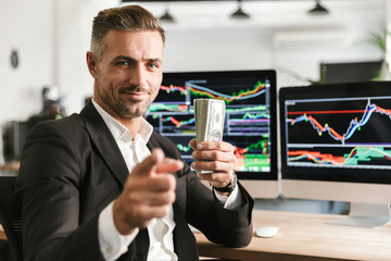 Fototapete - Image of handsome businessman holding pack of money while working in office with graphics and charts on computer