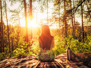 Beautiful young girl sitting on a plaid in a forest glade during sunset bright sunlight around beauty of nature. Next to the girl is an iron thermos with cups. Unity with nature concept. Girl enjoying