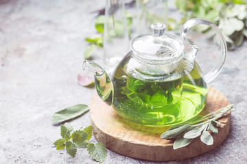 Hot herbal sedative mint tea drink in a glass teapot on wooden tray with fresh garden mint and sage leaves over gray background