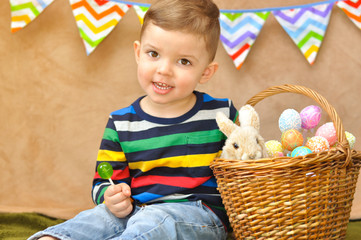 A little cute boy in a bright striped raglan sits near with a basket in which are Easter eggs and a little plush rabbit. A boy eats a green lollipop.