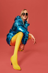 Young confident blonde girl in blue jacket squats in studio before camera, red background