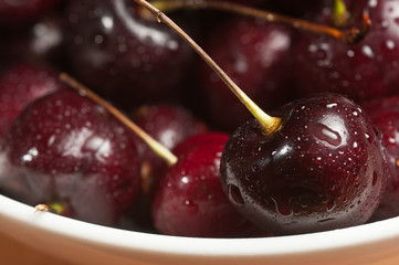 Top, front view , close distance of a white ceramic bowl of ripe, local, raw cherries with stems, rinsed on  white ceramic saucer, on a wood tabl