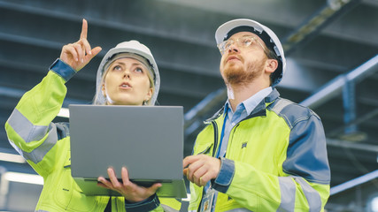 Fototapete - Male and Female Industrial Engineers Work on a Manufacturing Plant, They Discuss Project, Point in the Direction of the Machinery while Using Laptop. In the Background Industrial Manufacturing Plant.