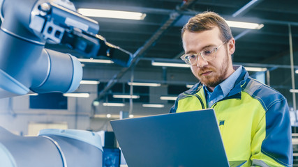 Fototapete - At the Factory: Automation Engineer Uses Laptop for Programming Robotic Arm. New Era in Automatic Manufacturing Industry.
