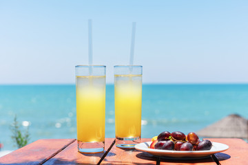 Two glasses of fresh tropical fruit juice on the beach, and a plate with grapes and watermelon on the table, against the sea. Holiday and vacation concept