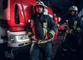 Wall Mural - Two firemen wearing protective uniform standing next to a fire engine in a garage of a fire department.