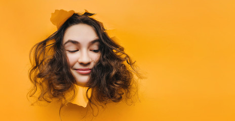 Wall Mural - A funny teenage caucasian girl with problematic dry curly hair expresses displeasure on her face. Orange, yellow paper background. Smile, grin.