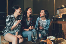 Group Of Young People In Rooftop Party Talking Sitting On Wooden Chairs. Happy Girls Enjoy Barbecue At Night On The Roof. Full Of Alcohol Drinks And Food On The Table Outdoors.