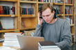 © taisiyakozorez - A young student man in headphones listens to music, communicates in a headset, works on a computer. Coworking in the library on the background of the bookshelf.