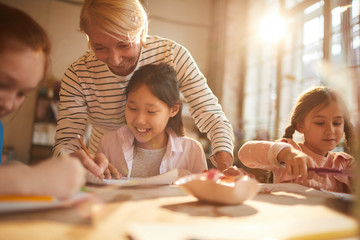 Wall Mural - Portrait of woman working with group of little girls painting in art class, scene lit by serene sunlight, copy space