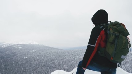 Wall Mural - A young man, a tourist with a backpack, stands on the edge of a mountain next to a cliff and admires the top of a mountain. Winter season. Slow motion.