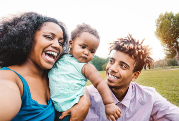 Happy African family doing selfie photo with mobile phone in a public park outdoor - Mother and father having fun with their daughter during a weekend sunny day - Love and happiness concept