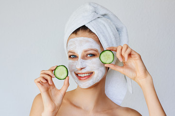 Beautiful young woman with facial mask on her face holding slices of cucumber. Skin care and treatment, spa, natural beauty and cosmetology concept.