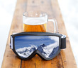Ski goggles with reflection of mountains and glass mug with fresh cold beer