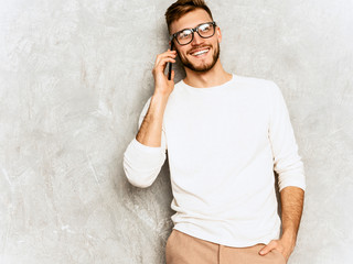 Portrait of handsome smiling hipster lumbersexual businessman model wearing casual summer white clothes. Fashion stylish man posing against gray wall. Speaking on mobile phone in spectacles