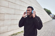 © guas - urban happy smiling african man in wireless headphones enjoying listening to music wearing black hoodie on city street over gray wall background