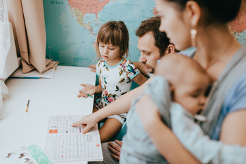 Canvas Print - Mother holds a baby while father helps his little daughter to make a lessons at the desk in the room with the map on the wall