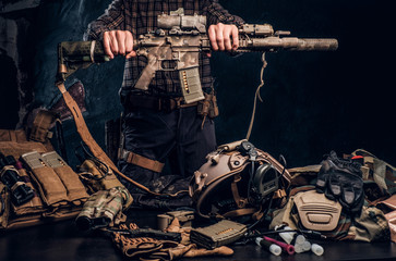 Wall Mural - Man in a checkered shirt holding a assault rifle and showing his military uniform and equipment. Modern special forces equipment. Studio photo against a dark textured wall