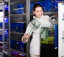 Wall Mural - Girl holding aquarian net and water container next to aquarium with colorful fish in aquarium shop