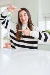 Wall Mural - Beautiful young woman wearing stripes sweater smiling making frame with hands and fingers with happy face. Creativity and photography concept.