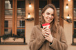 © bodnarphoto - Happy girl stands in the street with a cup of coffee in her hands, looks at the camera, smiles, poses on the camera.Smiling girl in warm clothes walking along the streets of the town, drinking coffee.