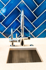 Wall Mural - New small modern faucet and kitchen sink closeup with countertop, blue vibrant backsplash and shiny clean stainless steel handle