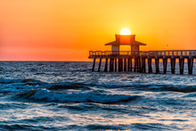 Naples, Florida Yellow And Orange Red Sunset Closeup In Gulf Of Mexico With Sun Inside Pier Wooden Jetty, And Many Birds Silhouette Flying Over Horizon And Blue Ocean Waves