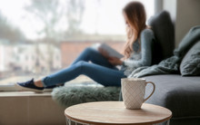 Cup On Table Of Young Woman Reading Book On Window Sill