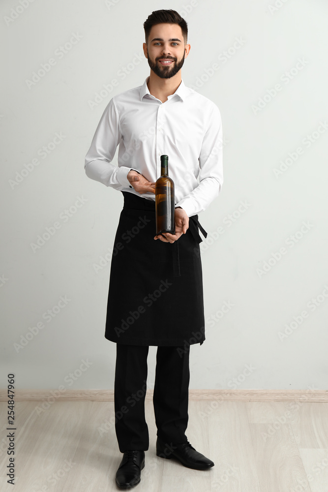 Handsome waiter with bottle of wine near light wall