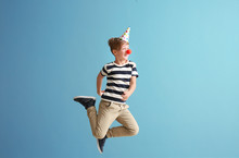 Jumping Little Boy With Clown Nose And Party Hat Against Color Wall. April Fools' Day Celebration
