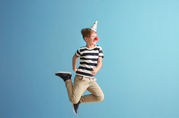 Jumping little boy with clown nose and party hat against color wall. April fools' day celebration