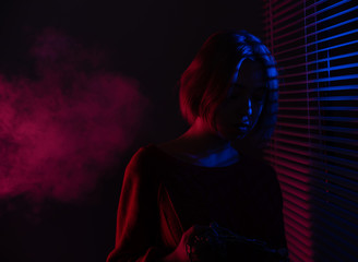 A beautiful girl with a chain in her hands stands near the window with blinds in the light of neon lights of blue and red light