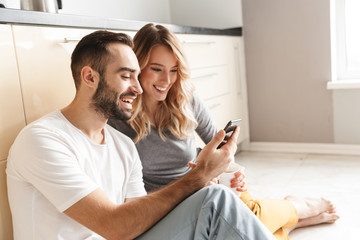 Poster - Amazing young loving couple sitting at the kitchen using mobile phone on a floor.