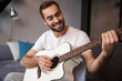 © Drobot Dean - Photo of attractive man playing acoustic guitar while sitting on sofa in apartment