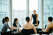 © Danon - A young Caucasian professional woman is leading a meeting with her team in a meeting room. She is professionally turned out in a black top and is professionally looking as she speaks to her team.