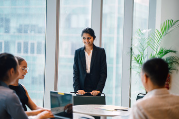 Wall Mural - An Asian Indian businesswoman is having a conversation with her colleagues in a conference room. She's smiling elegantly.  She's wearing a black blazer and white blouse with a black skirt.